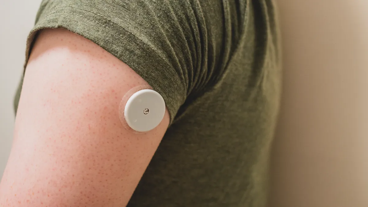 A round white medical sensor installed on the back of the upper arm. The sensor is descrete even though it’s installed just below the T-shirt line. The sensor is about the size of a chunky…