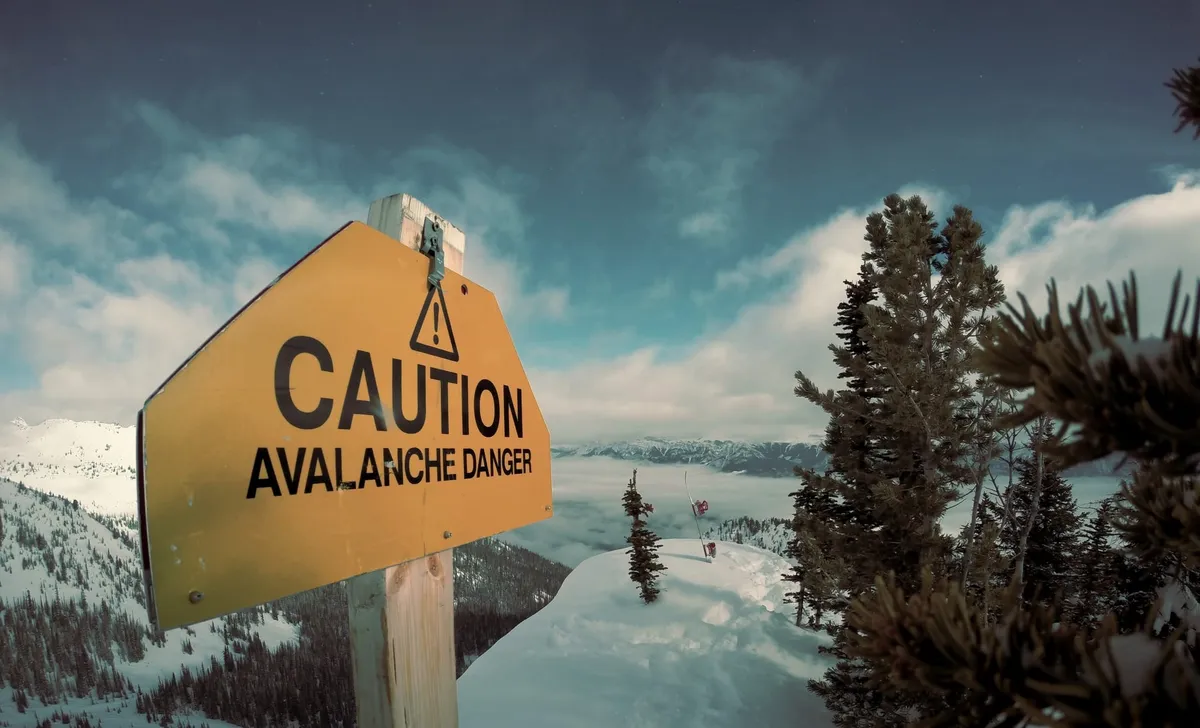 A warning sign saying “Caution! Avalanche danger.” with a snow-covered hill in the background.