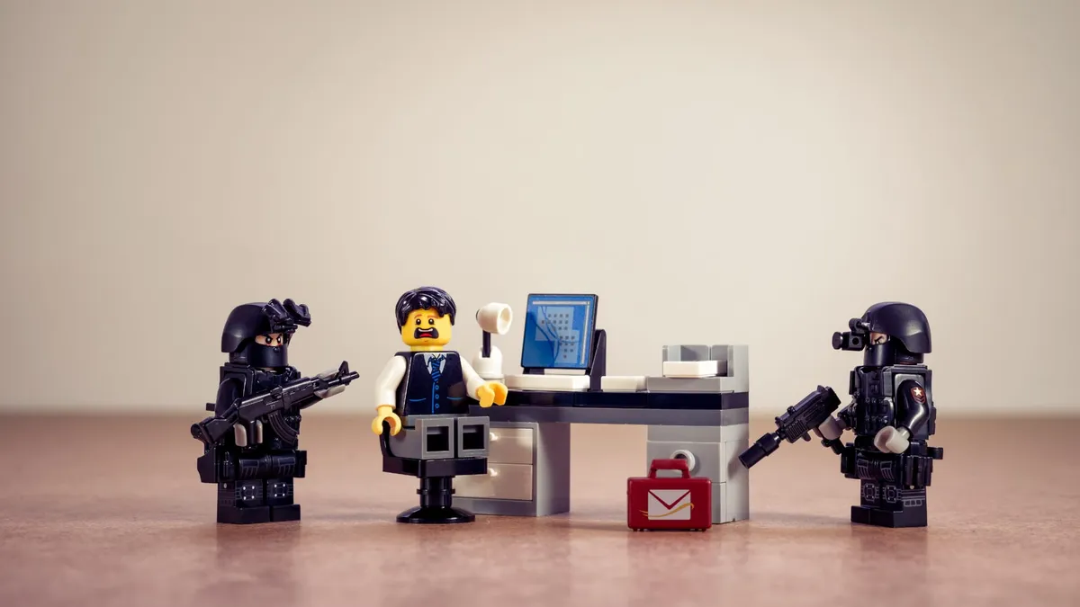 A Lego minifigure set with one scared looking figure sitting at a desk in front of a computer surrounded by two minifigures holding machine guns and wearing tactical armor.