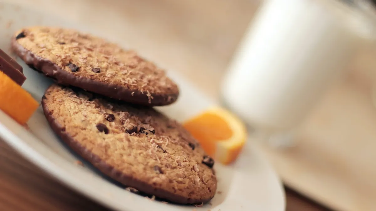 Two cookies on a plate with some oranges and a glass of milk in the background.