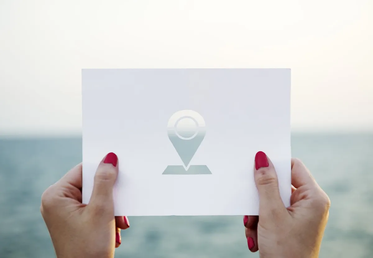 A pair of hands holding up a paper card with a geolocation marker icon cut out of the paper.