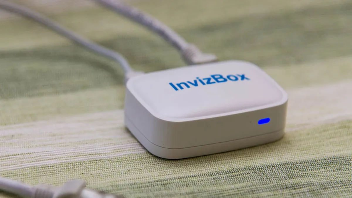 A small white box labeled “Invizbox” sitting on a table with a blue status light on the front and two cables sticking out on the back.