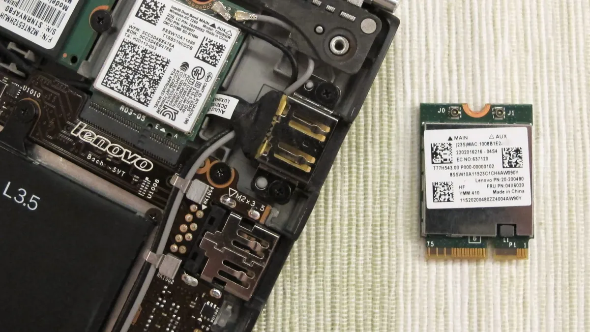 A laptop with the back cover removed and the Wi-Fi module laying next to it on a table.
