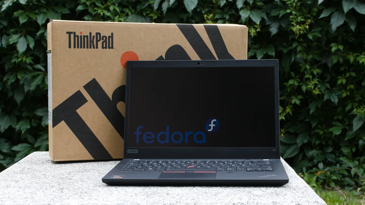 A Lenovo ThinkPad T14 laptop open with the Fedora Linux logo shown on screen. The original packaging is shown behind it.