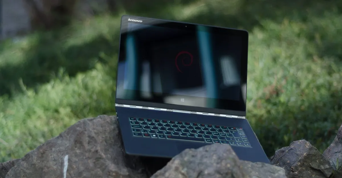 A Lenovo Yoga 3 Pro laptop sitting on a rok outdoors with grass in the background. The Debian logo is shown on screen.