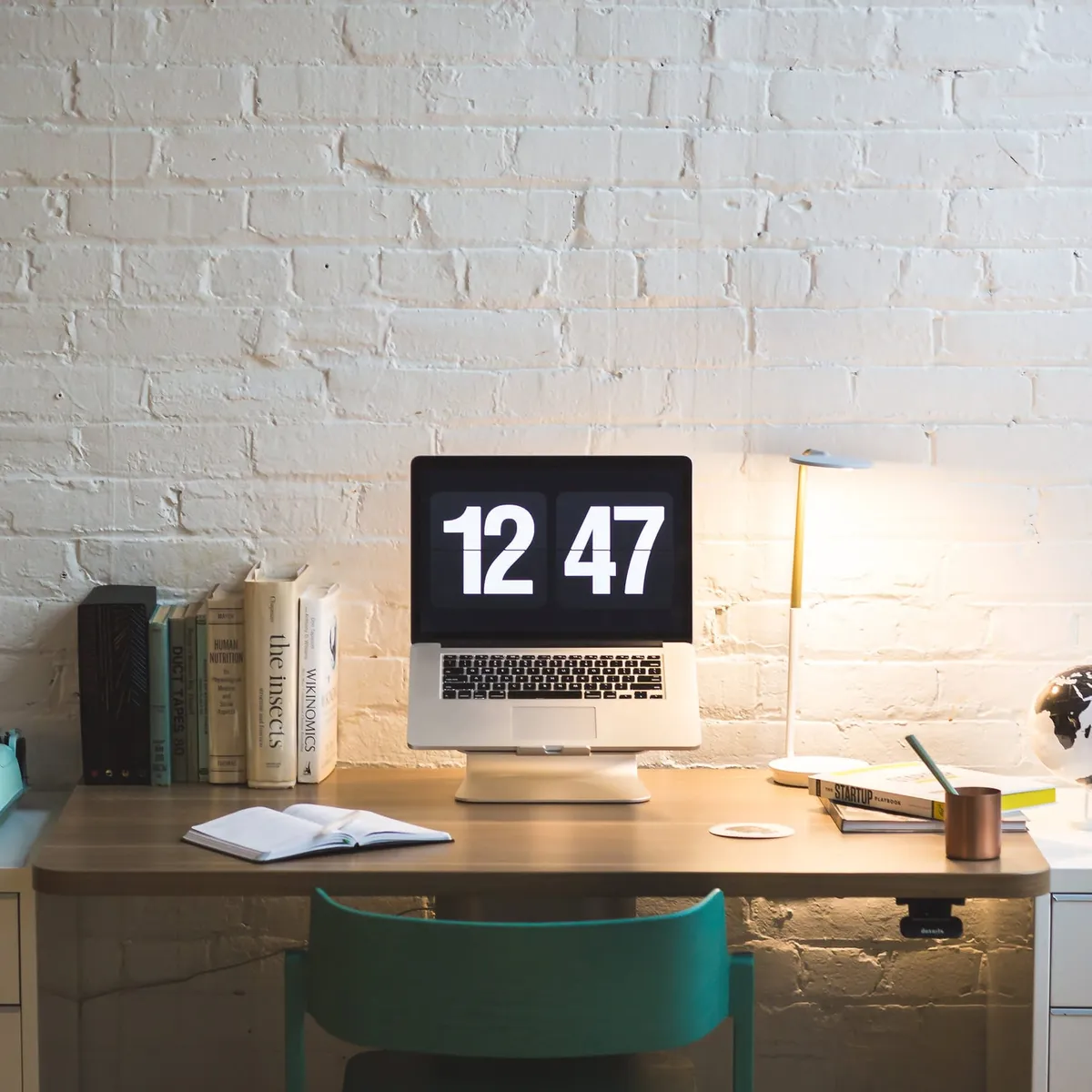 A MacBook laptop on a cluttered desk. The screen is showing a large digital clock at 12:47.