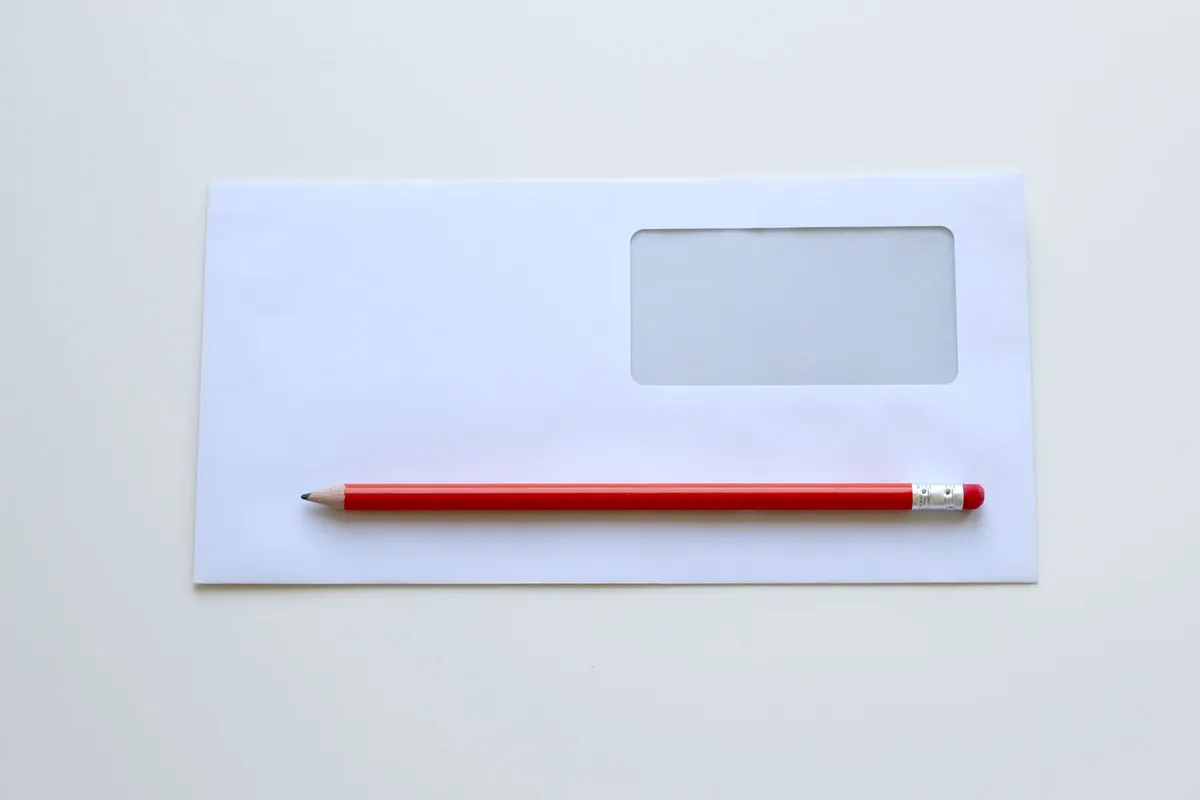 A red pencil laying on top of an envelope laying on a table.