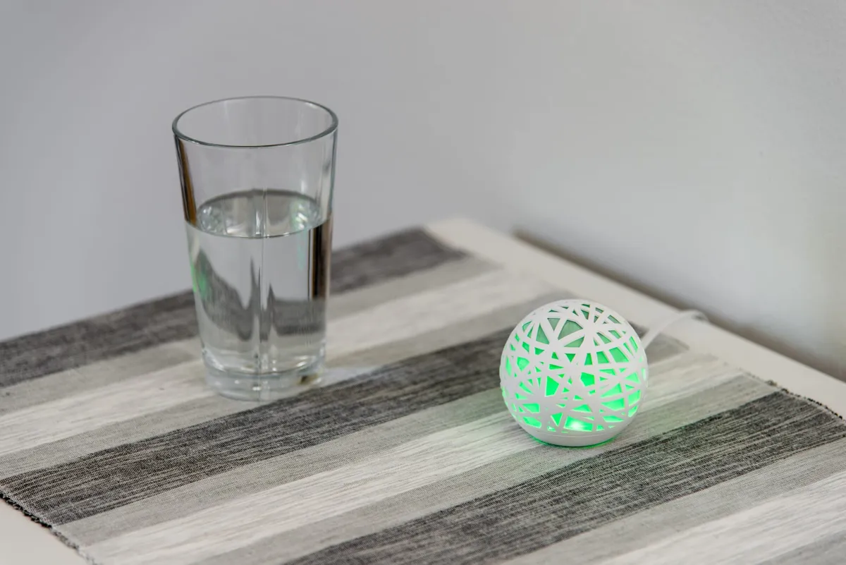 A green glowing Sense sleep monitoring device sitting on a bedside table next to a glass of water.