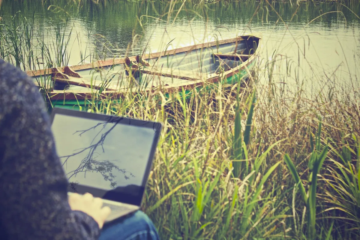 A person working outside on a laptop with a rowboat in the background.