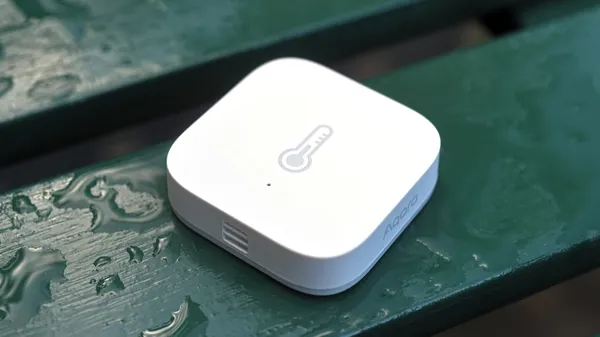 An white and compact Aqara Temperature and Humidity sensor laying on a rain wet bench.