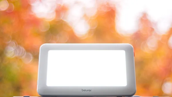 A lit Beurer TL20 daylight therapy lamp against a backdrop of autumn leaves.