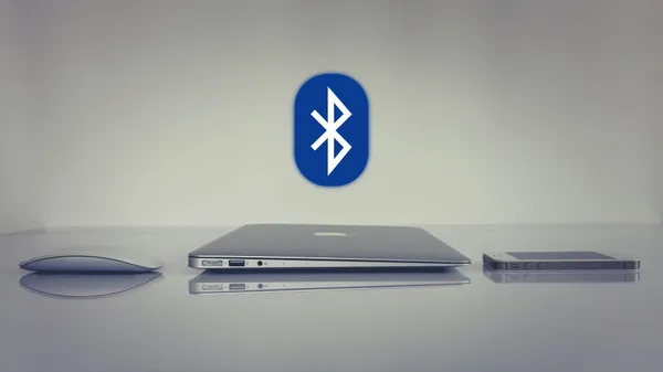A computer mouse laying next to a laptop and a phone with the Bluetooth icon looming in the background.