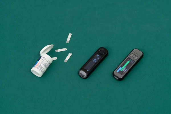 An open box of glucose test strips spills out on a green surface. Two different glucose meters sits next to it.