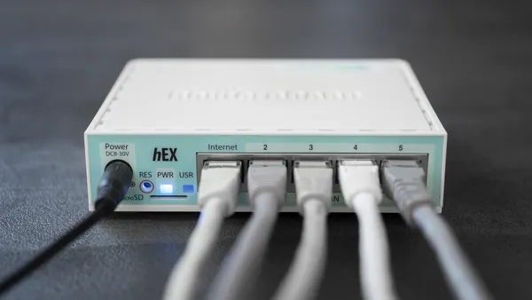A MikroTik RouterBOARD RB750Gr3 network router with five connected Ethernet cables. One Ethernet port is labelled “Internet.”