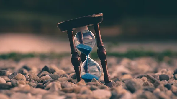 An hourglass with blue sand halfway through its countdown is resting on some rocks.