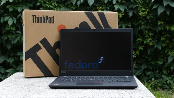 A Lenovo ThinkPad T14 laptop open with the Fedora Linux logo shown on screen. The original packaging is shown behind it.