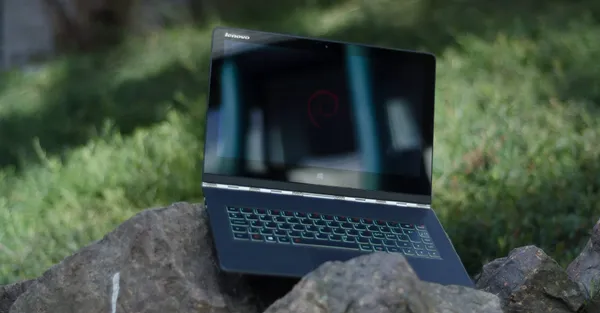 A Lenovo Yoga 3 Pro laptop sitting on a rok outdoors with grass in the background. The Debian logo is shown on screen.