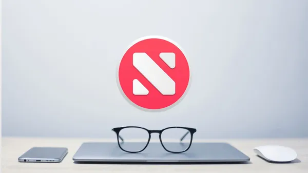 The Apple News logo hovering in mid-air above a MacBook with reading glasses on top of it. An iPhone and an Apple Mouse is next to the MacBook.