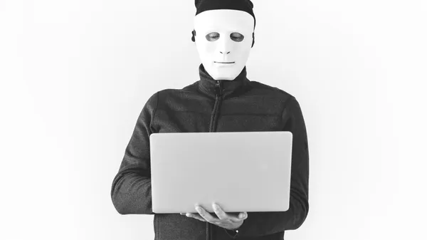 A man wearing a black padded jacket and a white mask covering his entire face holds a gray laptop computer.