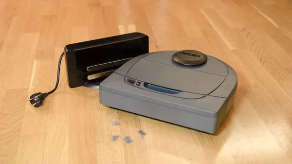 A Neato BotVac D3 robotic vacuum cleaner in front of some dust bunnies with its charging station.