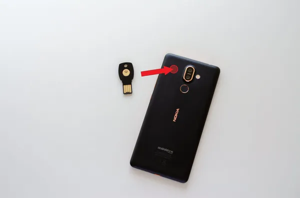 The backside of a Nokia 7 Plus smartphone with an arrow pointing from a blue NFC security key device and onto the left side of the camera on the smartphone.