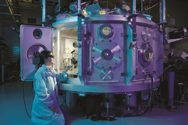 A female scientist wearing a lab coat and protective goggles looking into a big and bulky electronic device.