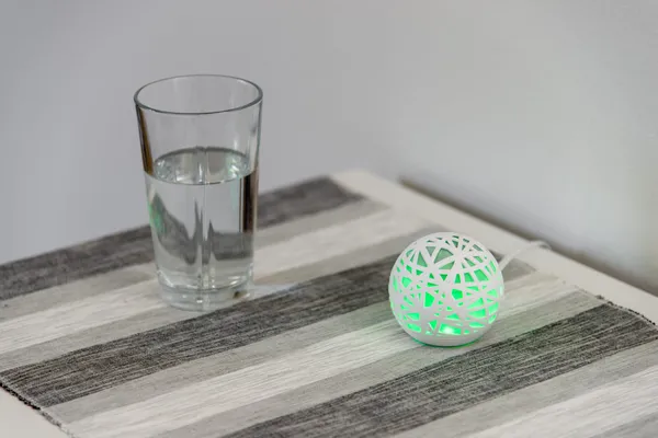 A green glowing Sense sleep monitoring device sitting on a bedside table next to a glass of water.