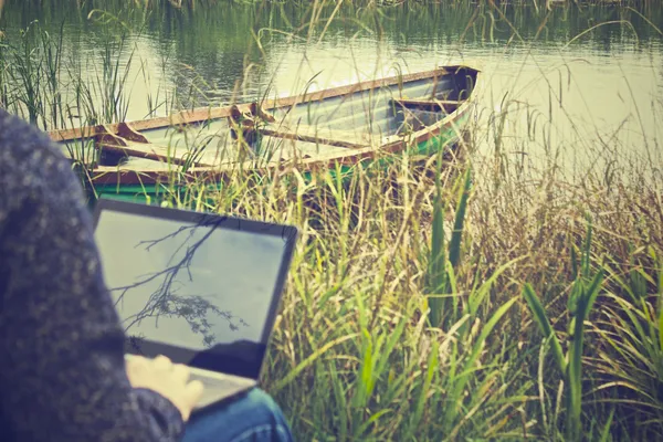 A person working outside on a laptop with a rowboat in the background.
