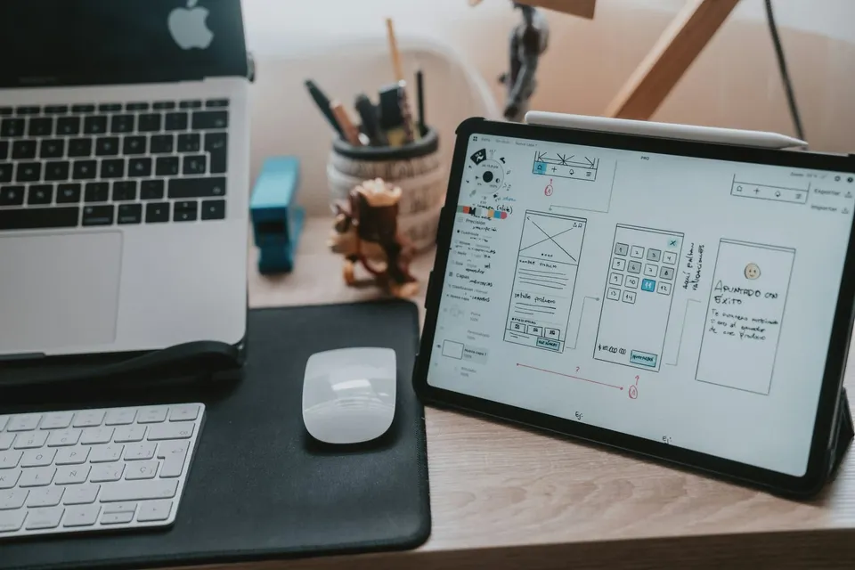 MacBook next to a tablet displaying a wireframe interface design on a desk.