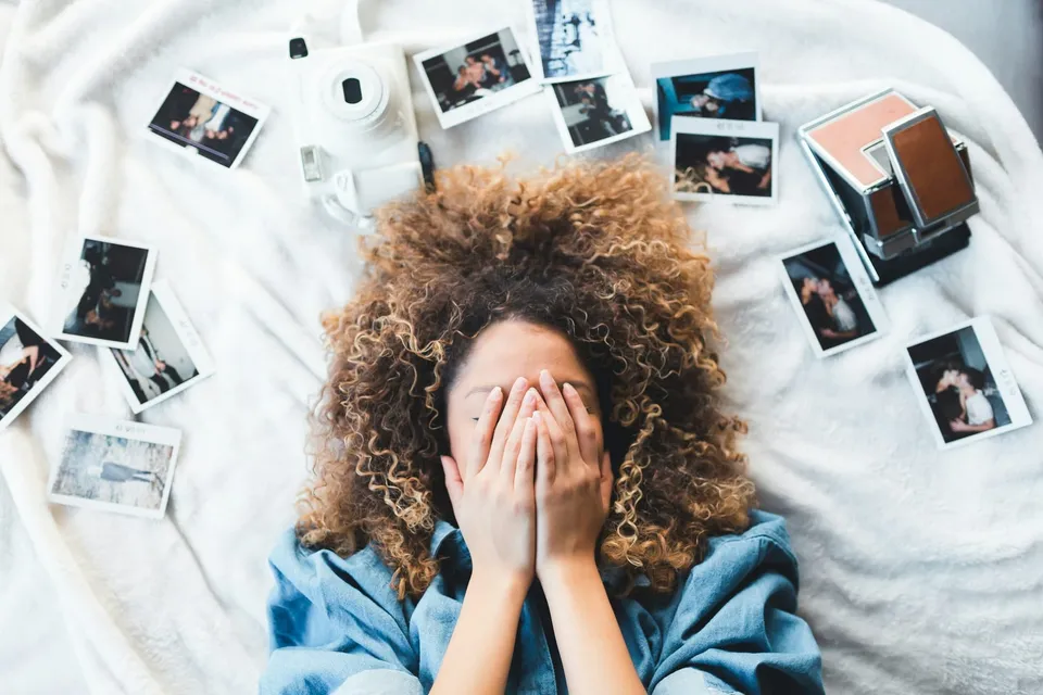 Woman lying on a bed with her face covered, surrounded by scattered photographs and a camera.