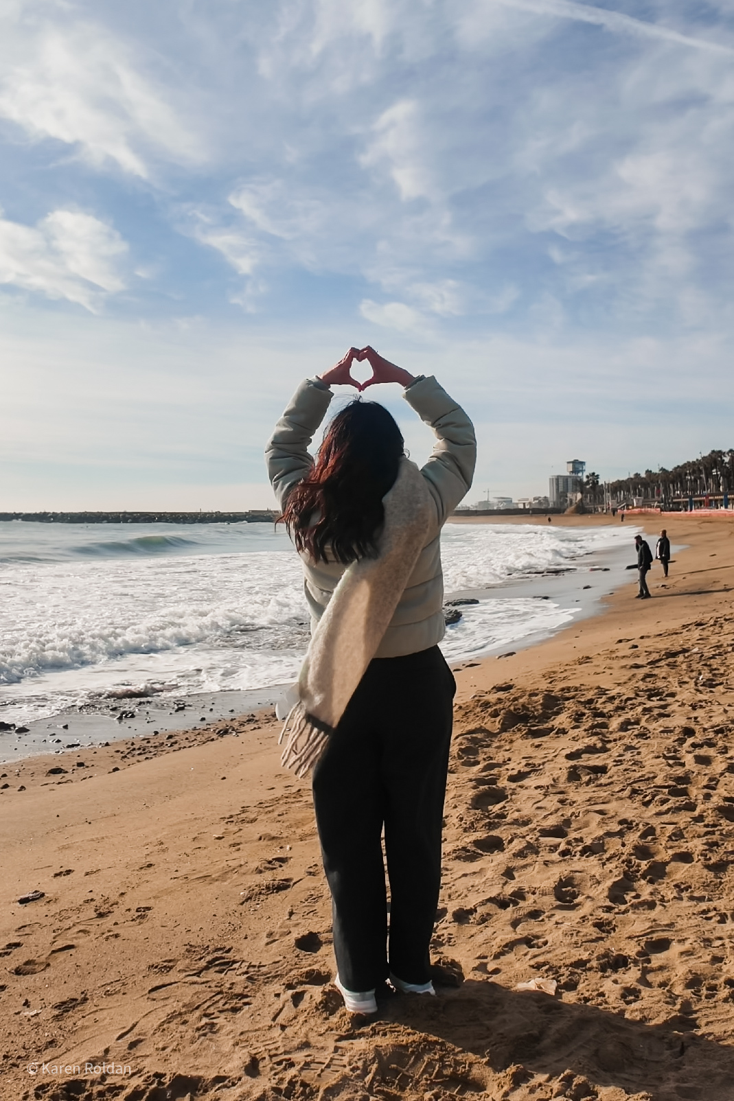 A woman making a heart shape with her hands while facing the ocean waves at Barceloneta Beach.