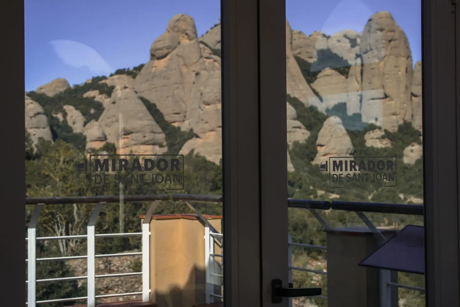 Panoramic view of rugged Montserrat mountains from the Mirador de Sant Joan viewpoint, with a balcony railing. Text: MIRADOR DE SANT JOAN.
