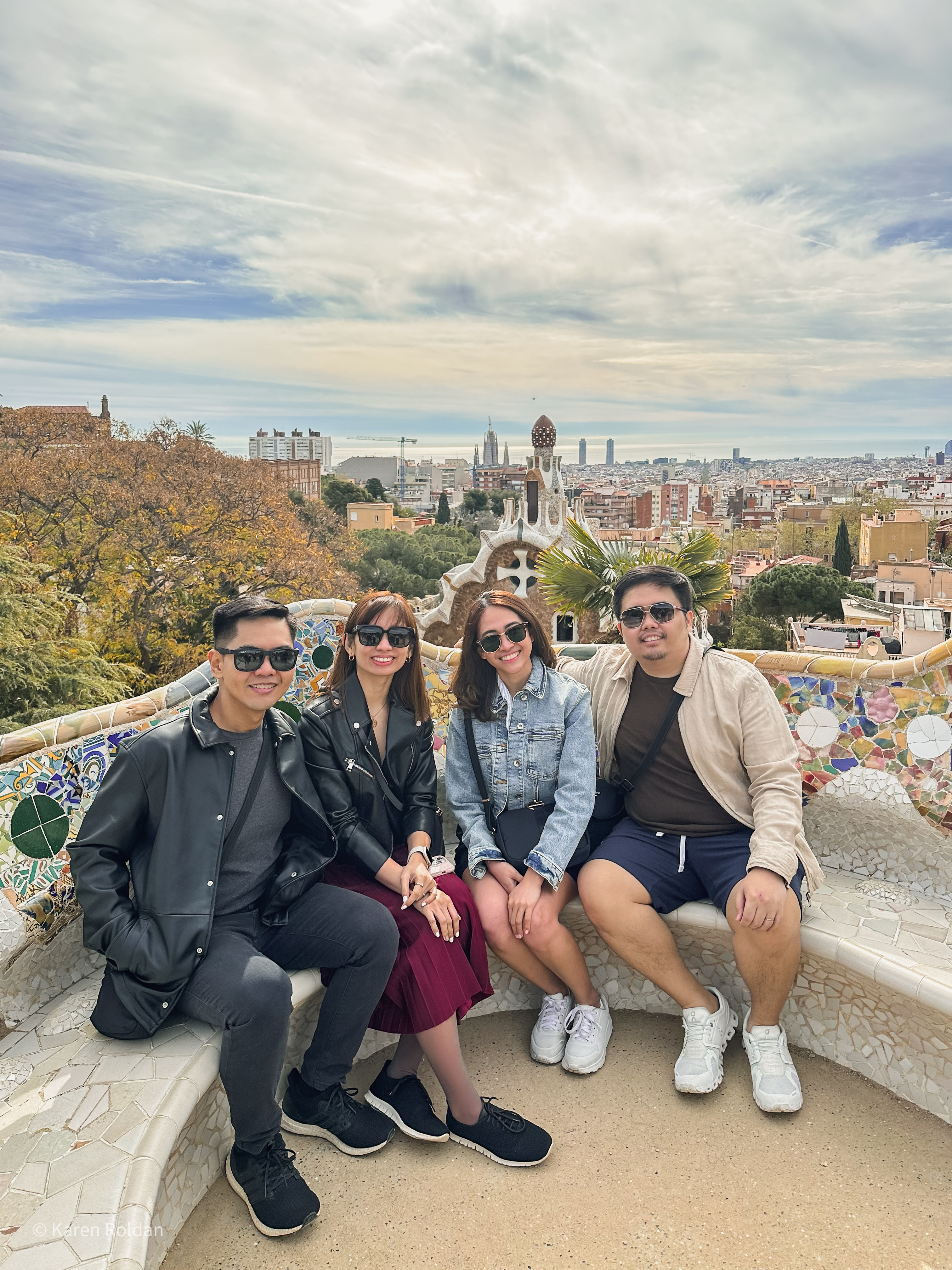 A group of friends sitting on the colorful mosaic Serpentine Bench at Park Güell with a panoramic view of Barcelona in the background.