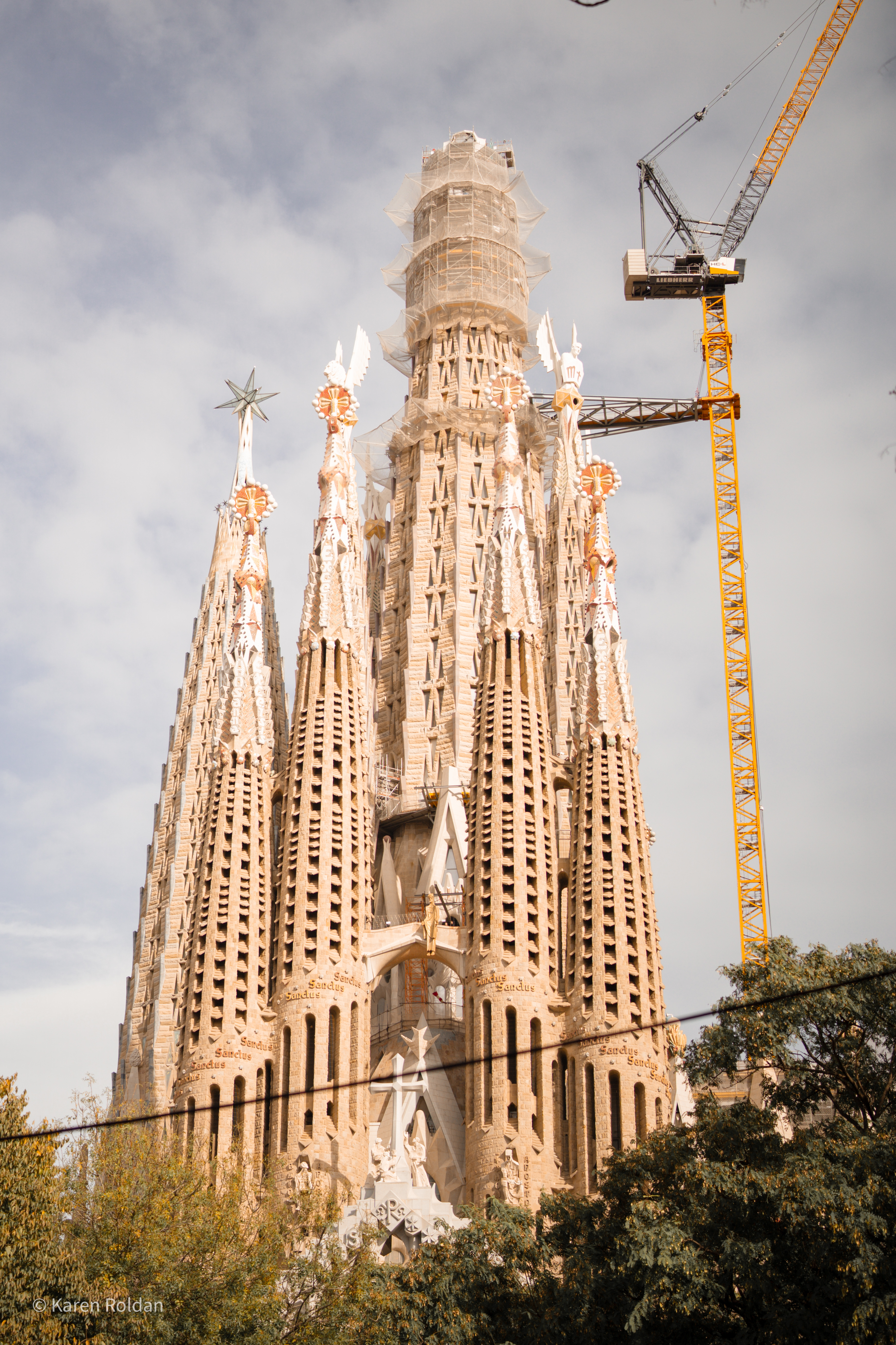 Low-angle view of the Sagrada Família's Passion Facade towers inscribed with 'Sanctus' and construction cranes.