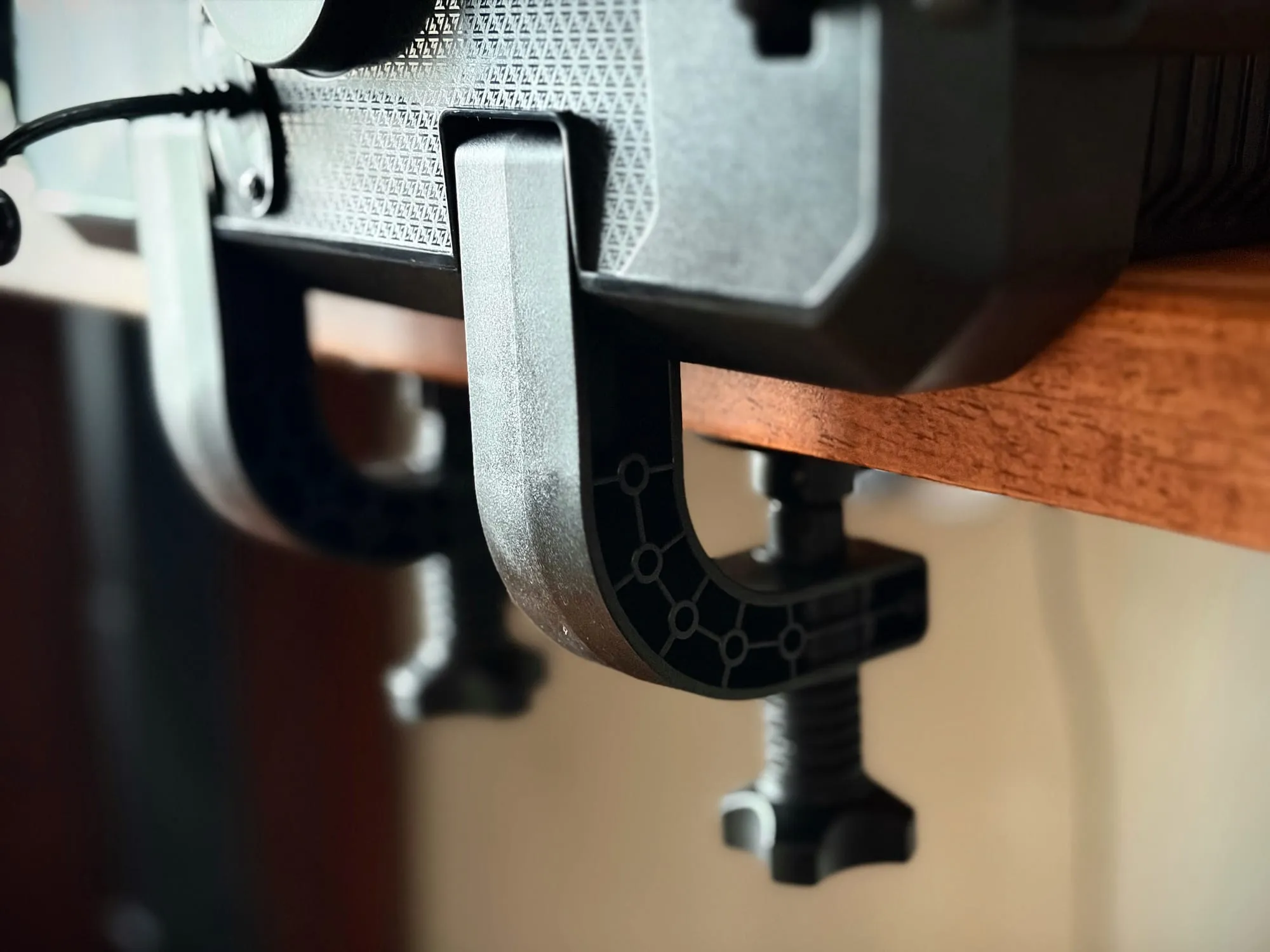 Two black plastic C-clamps gripping the edge of a light wood desk, each with a threaded screw and a plastic knob underneath.