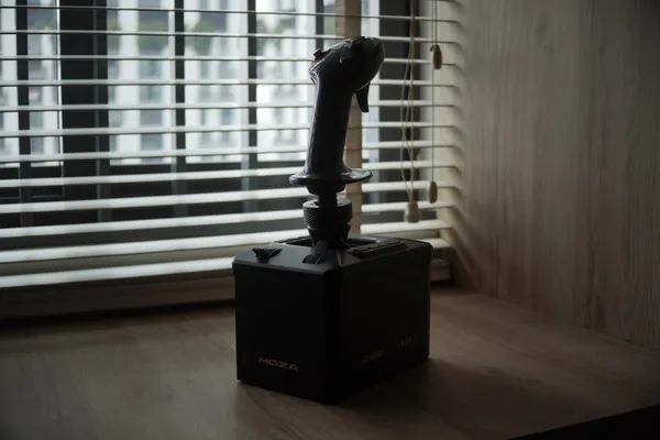 The MOZA AB6 force feedback base with a side stick grip fitted, standing on a wooden desk beside a window blind