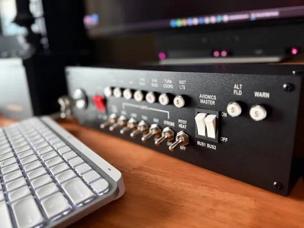 The Meza Aircraft Switch Panel on a wooden desk beside a keyboard: black faceplate, white legends, metal toggles for pitot heat and strobes, and the avionics rockers marked BUS1 and BUS2
