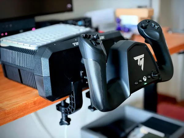 Thrustmaster TCA Sky Yoke clamped to the front edge of a light wood desk, a white keyboard resting on the flat top, monitor and speakers behind.
