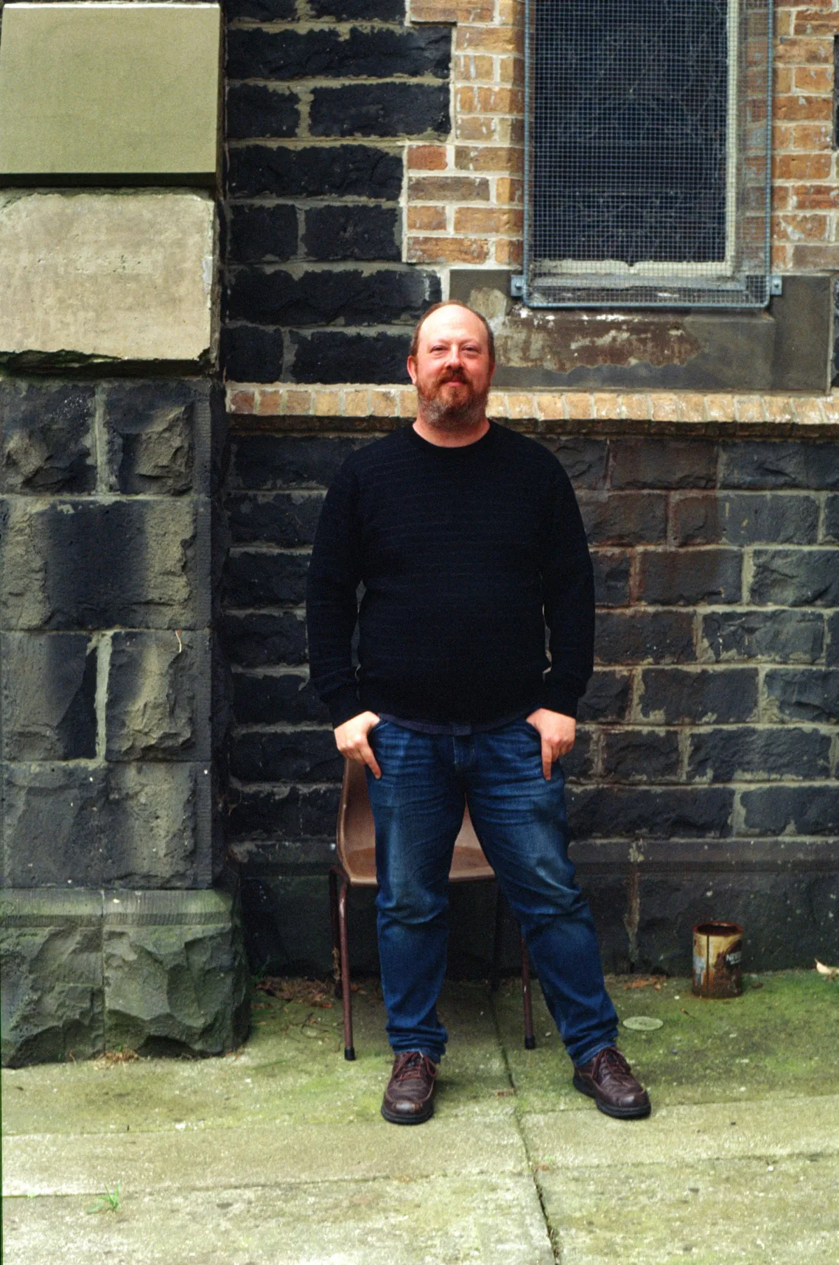An aging cartoonist wearing blue jeans and a dark jumper, standing in front of a church.