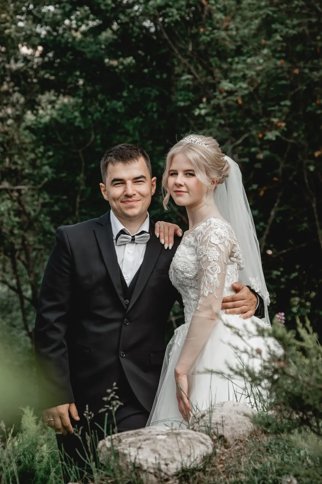 Couple in elegant wedding attire posing in a serene forest setting.