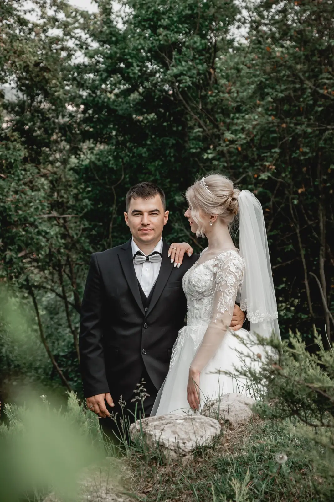 A couple poses outdoors in formal attire, with lush green trees in the background.