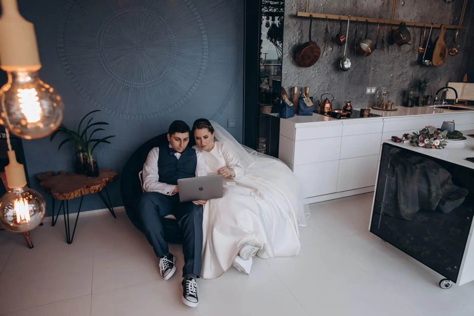 Bride and groom in kitchen reviewing wedding photos on a laptop, captured indoors.