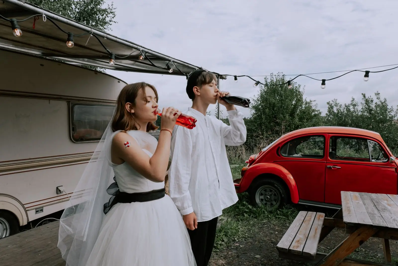 Couple in wedding attire enjoying drinks outdoors next to a retro car and camper.