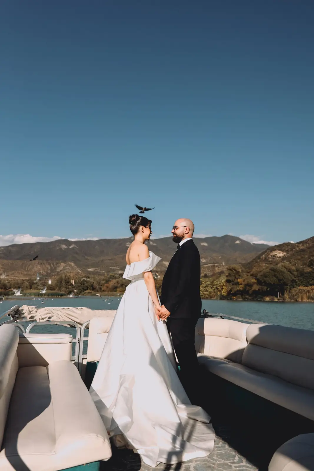 Bride and groom standing on a boat holding hands with mountains in the background.