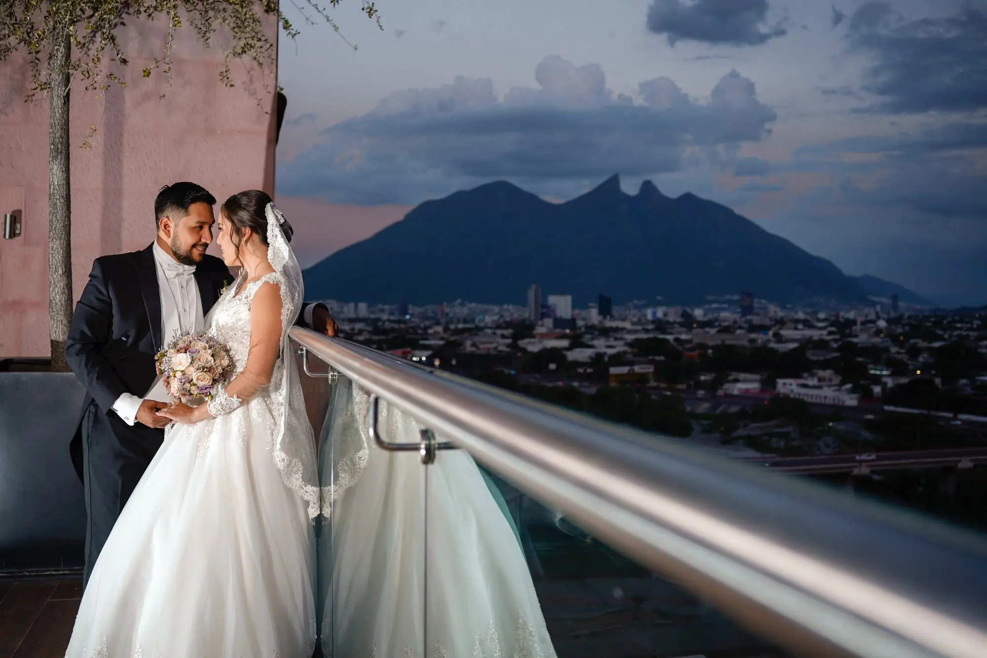 Bride and groom embrace lovingly on a balcony with stunning mountain backdrop at sunset.