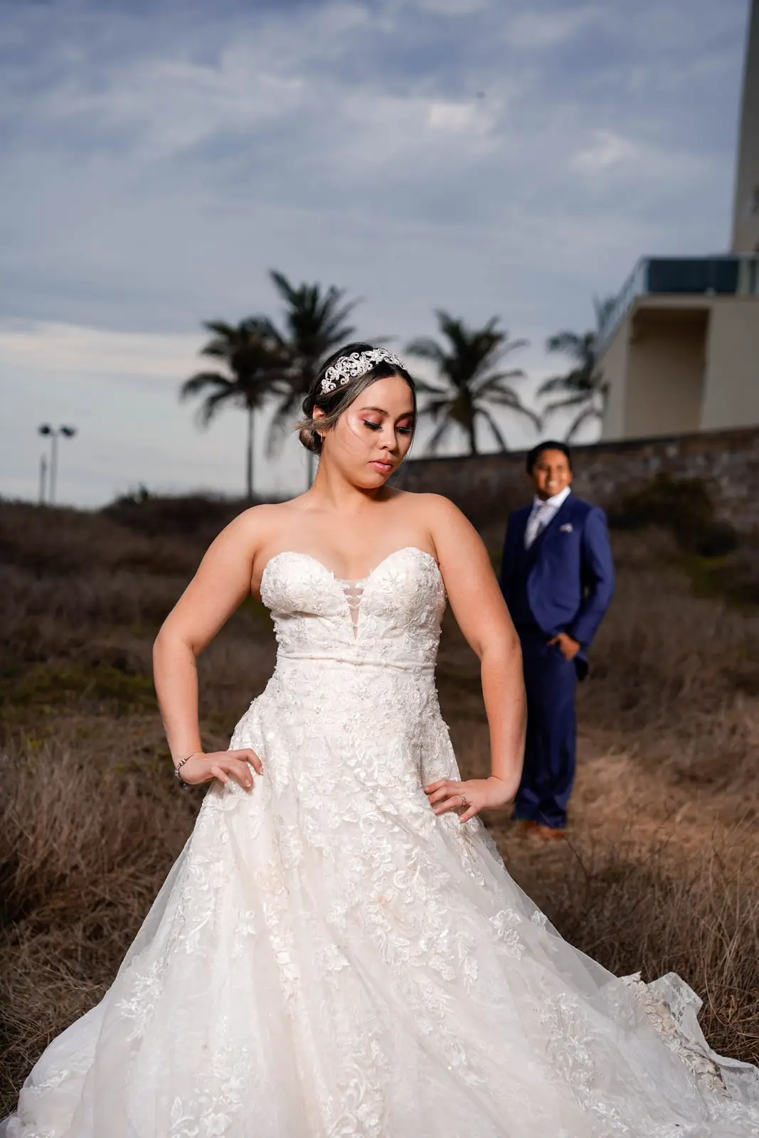 Bride in lace gown poses outdoors with groom in background during sunset.