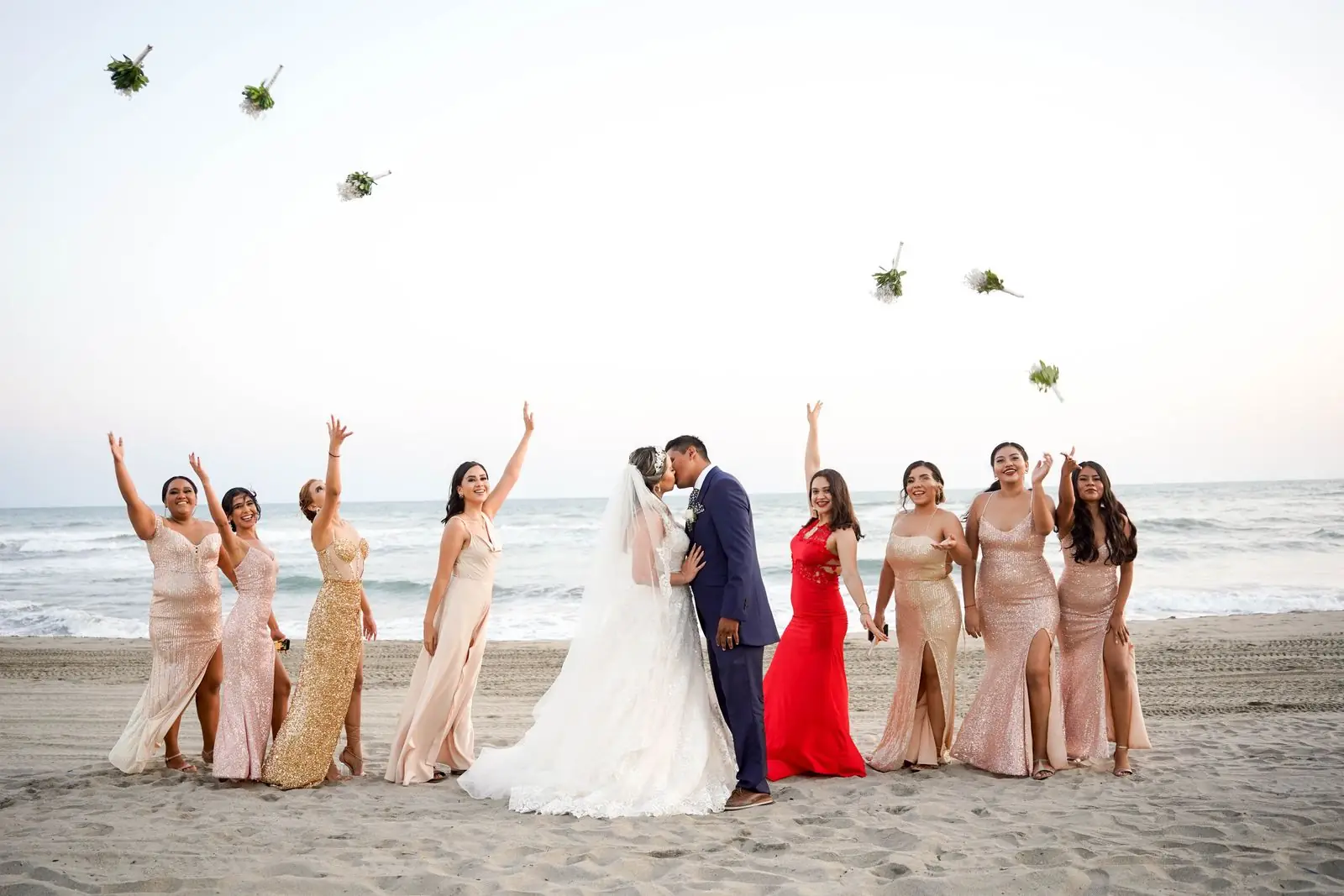 Bride and groom kissing on a beach surrounded by joyful bridesmaids during sunset.