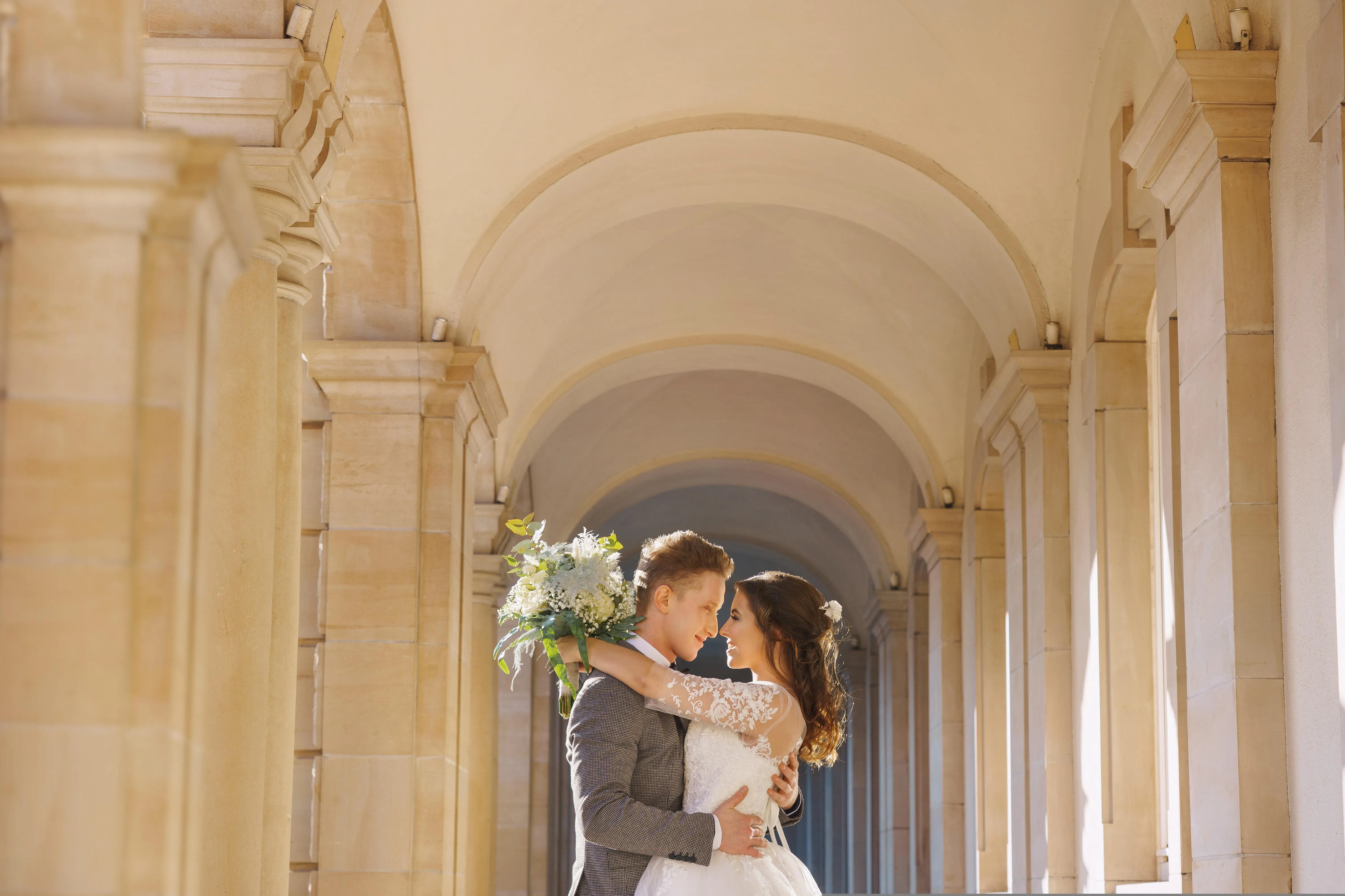 Bride and groom embracing beneath sunlit stone arches.