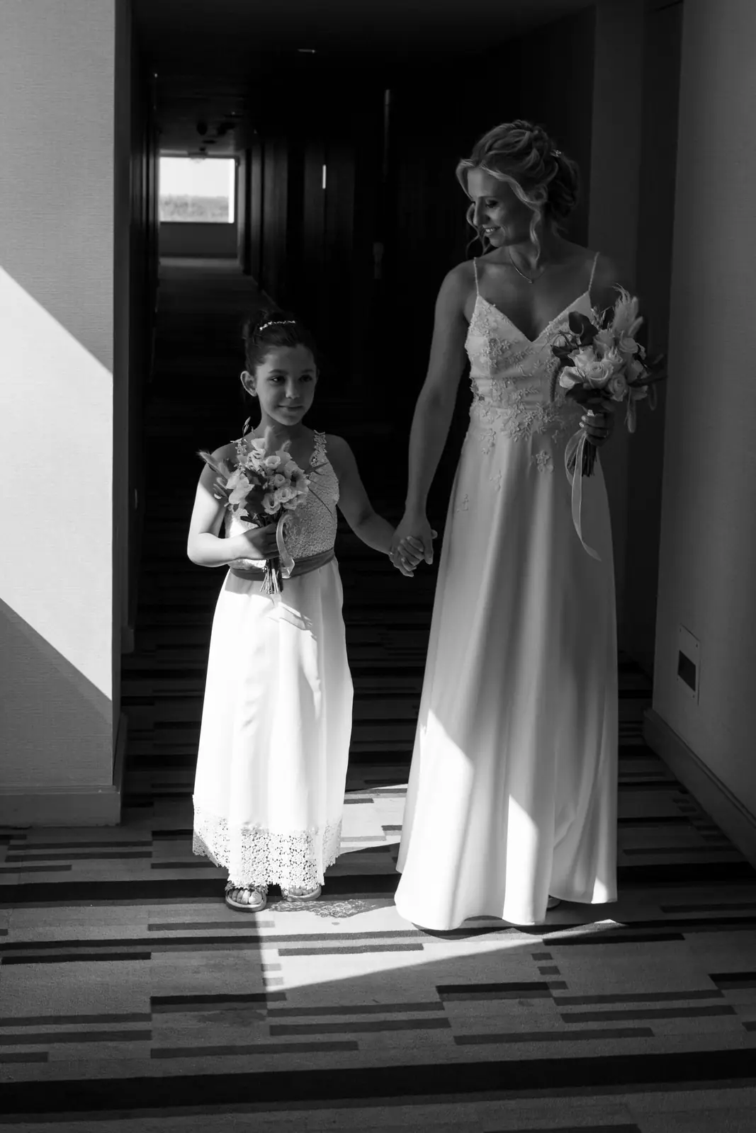 Black and white photo of a bride and child in wedding dresses holding flowers indoors.