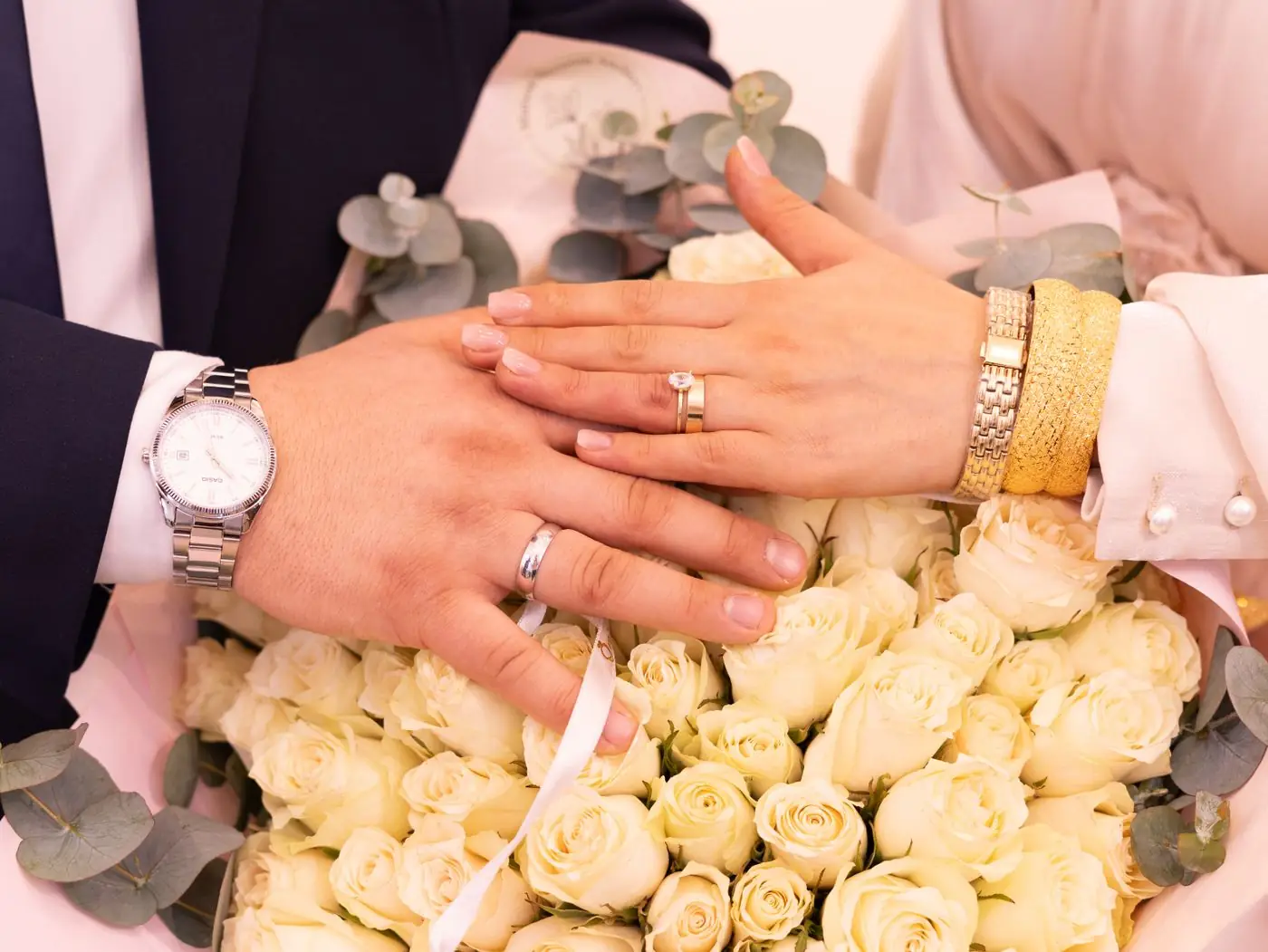 Close-up of wedding rings on a bride and groom's hands atop a bouquet of white roses.